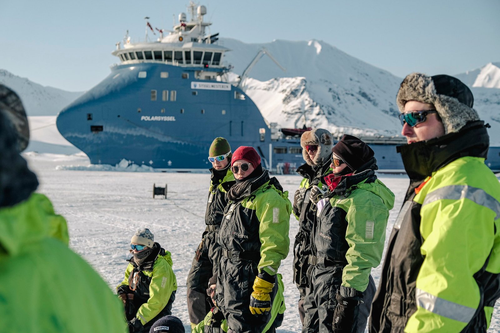Researchers in survival suits on sea ice in front of a research vessel